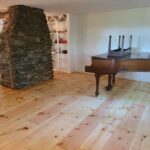 A room featuring a stone chimney and built-in shelves rests harmoniously on a wooden floor, the result of one of the most challenging sanding jobs. A grand piano completes the timeless elegance of this carefully crafted space.
