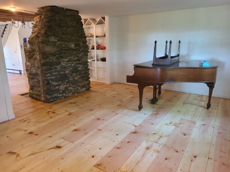A room featuring a stone chimney and built-in shelves rests harmoniously on a wooden floor, the result of one of the most challenging sanding jobs. A grand piano completes the timeless elegance of this carefully crafted space.