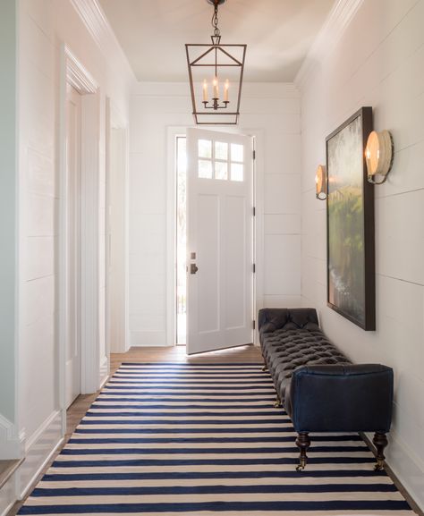 A hallway featuring hardwood floors adorned with a striped rug, a tufted bench, and a captivating painting. An open door reveals a warm glow from the lantern-style light fixture above.