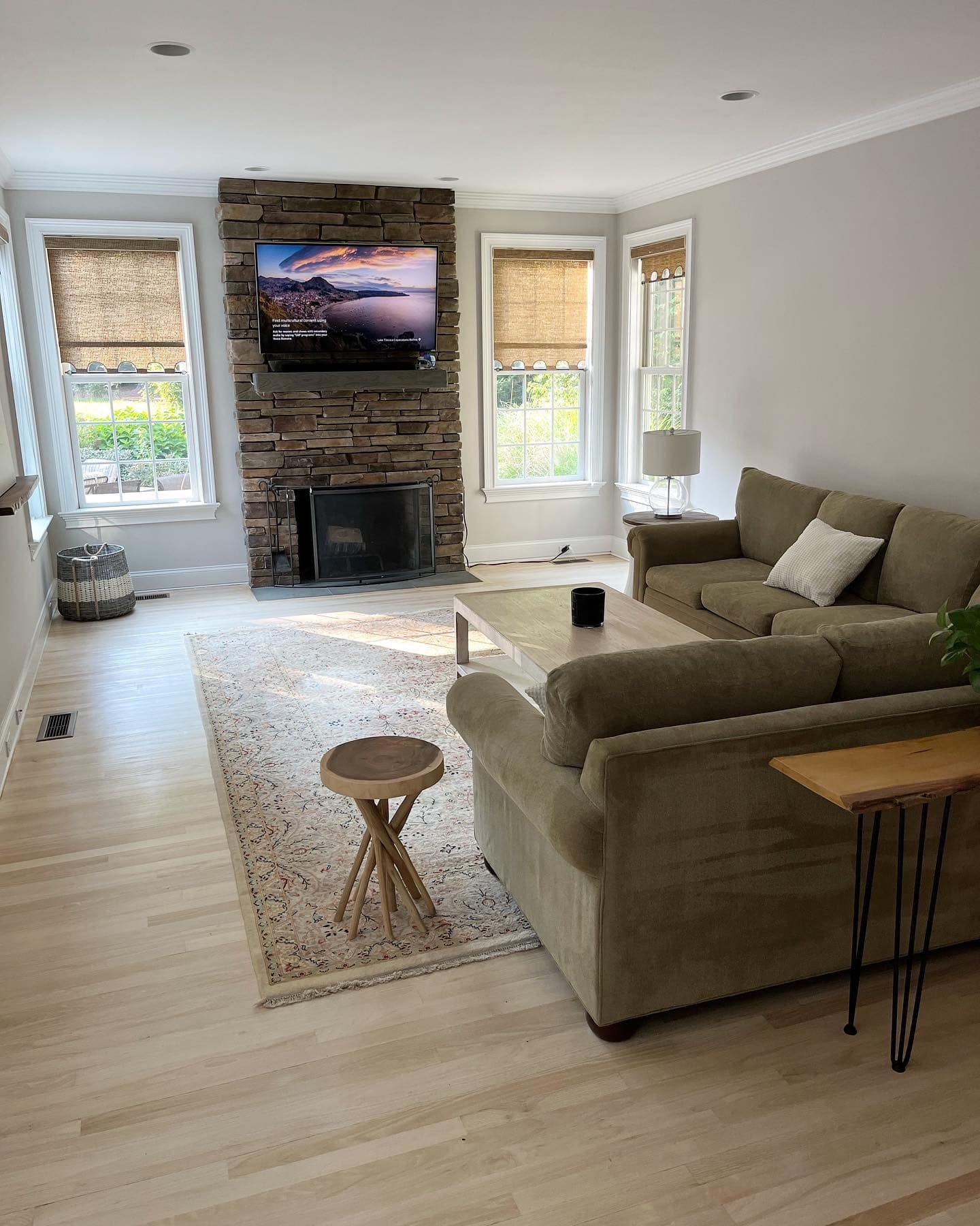 A cozy living room with a stone fireplace, wall-mounted TV, beige sofa, and large windows.