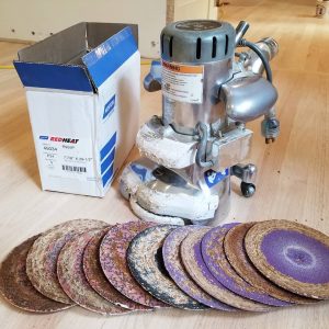 A floor sander sits next to a box and a row of various grit sanding discs on a wooden floor.