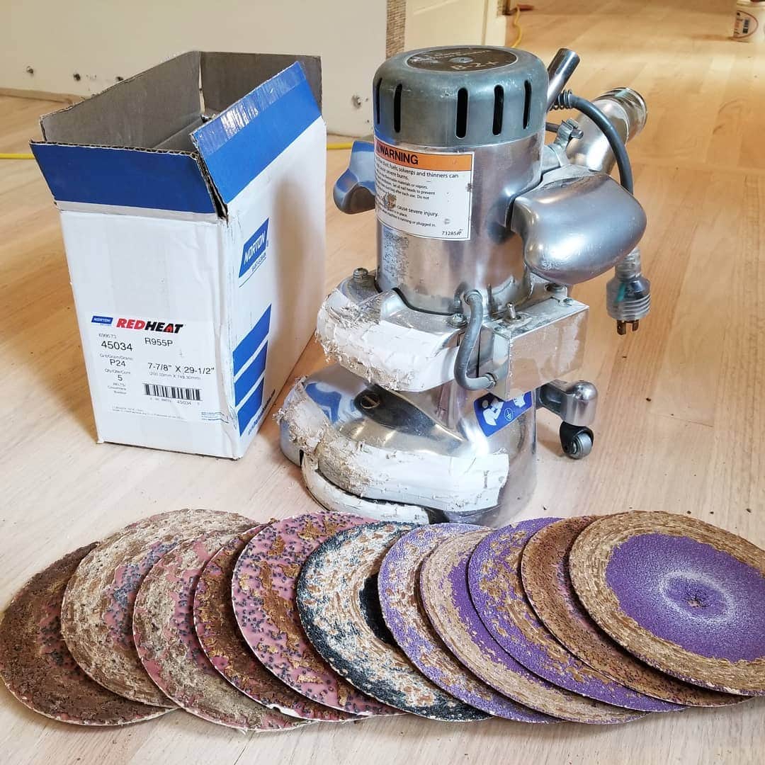 A floor sander sits next to a box and a row of various grit sanding discs on a wooden floor.