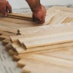 Hands installing wooden parquet flooring in a herringbone pattern on a concrete subfloor.
