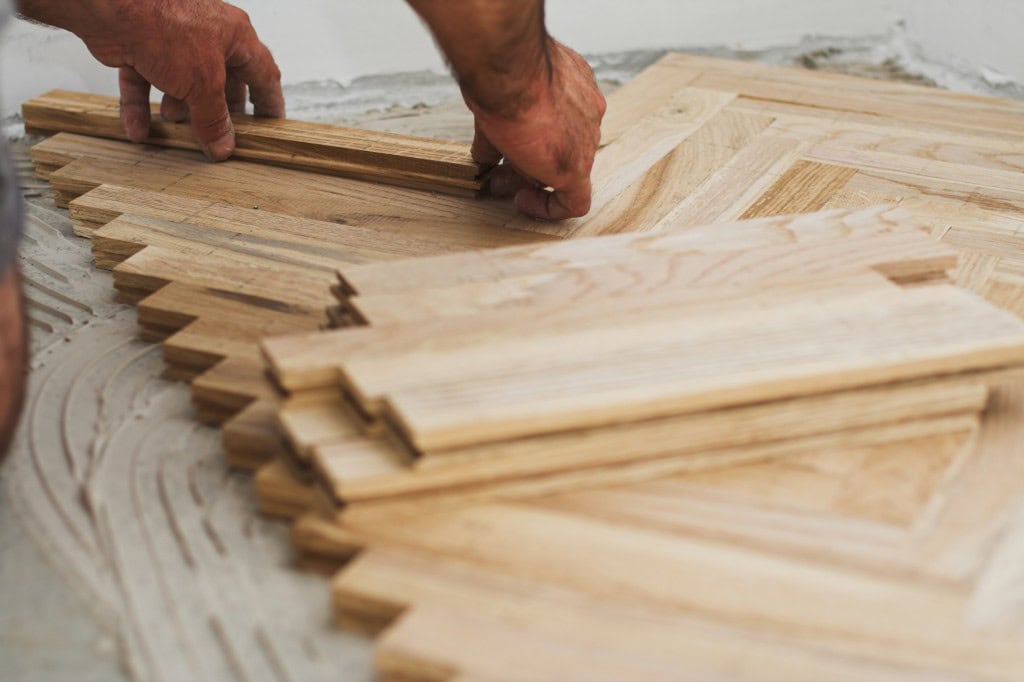 Hands installing wooden parquet flooring in a herringbone pattern on a concrete subfloor.