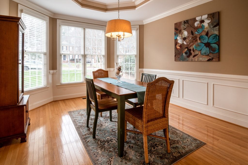 A bright dining room with a wooden table, four wicker chairs, and large windows letting in natural light.