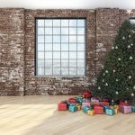 Christmas tree with ornaments and gifts beneath it in a loft with brick walls and large windows.