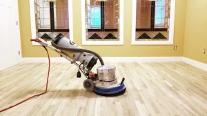 A floor sander sits on a light wood floor in an empty room with stained glass windows.