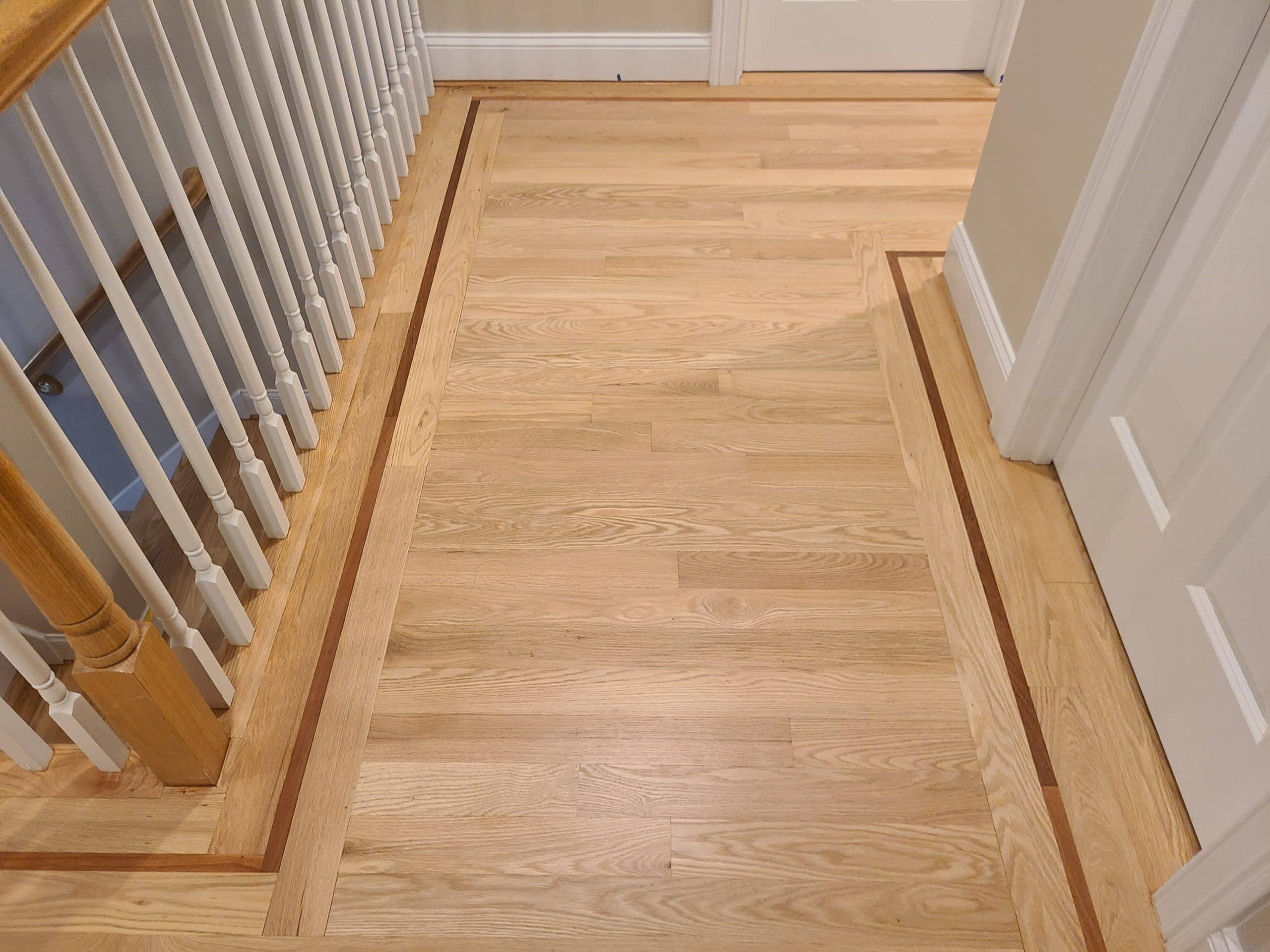 Light wood floor with a decorative border next to white doors and a staircase with white railings.
