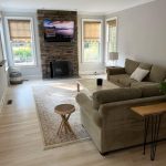 A cozy living room with a stone fireplace, wall-mounted TV, beige sofa, wooden coffee table, and large windows.