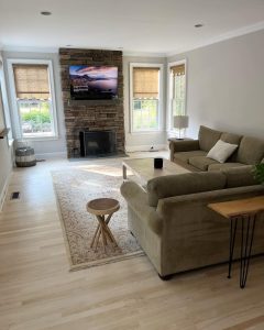 A cozy living room with a stone fireplace, wall-mounted TV, beige sofa, wooden coffee table, and large windows.