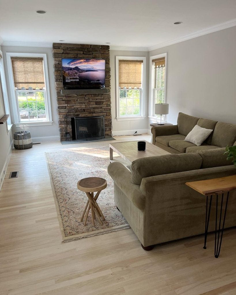 A cozy living room with a stone fireplace, wall-mounted TV, beige sofa, wooden coffee table, and large windows.